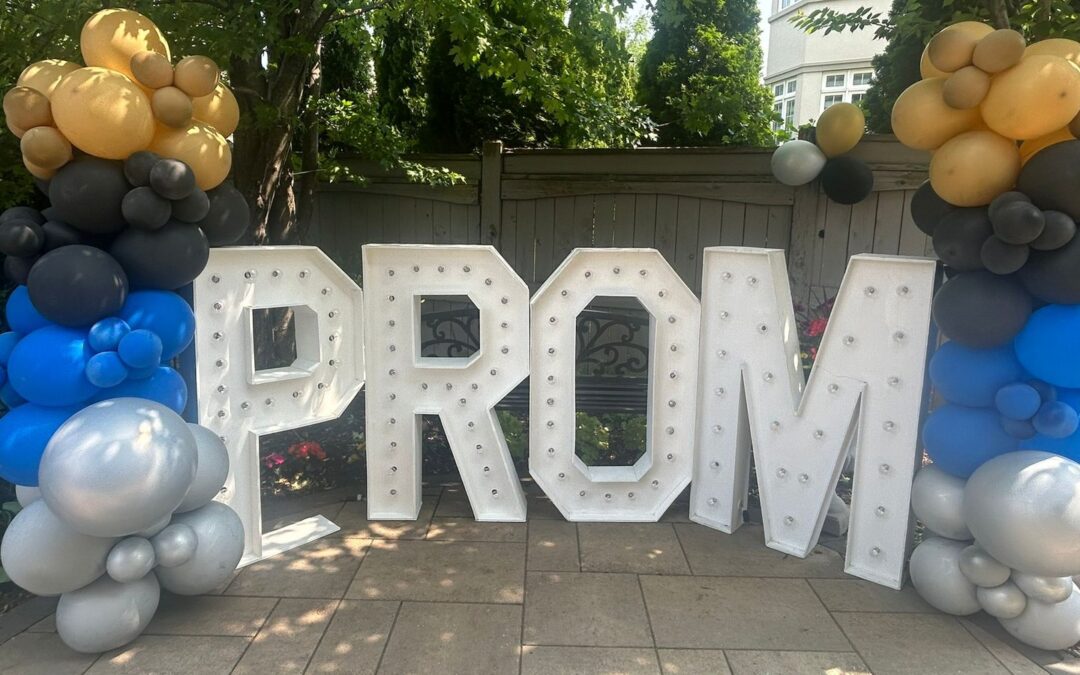 A pair of blue, silver, charcoal and gold balloon columns standing either side of large lit letters spelling PROM