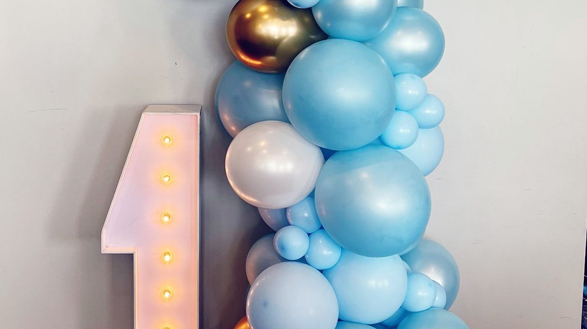 A blue, white and chrome gold balloon garland against a pale wall beside a lit marquee number