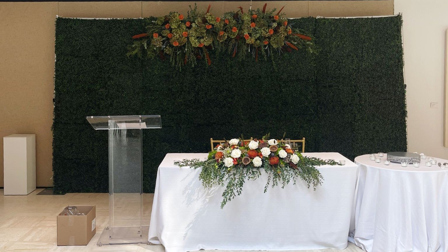 A green boxwood hedge wall with a floral swag standing behind a long white head table