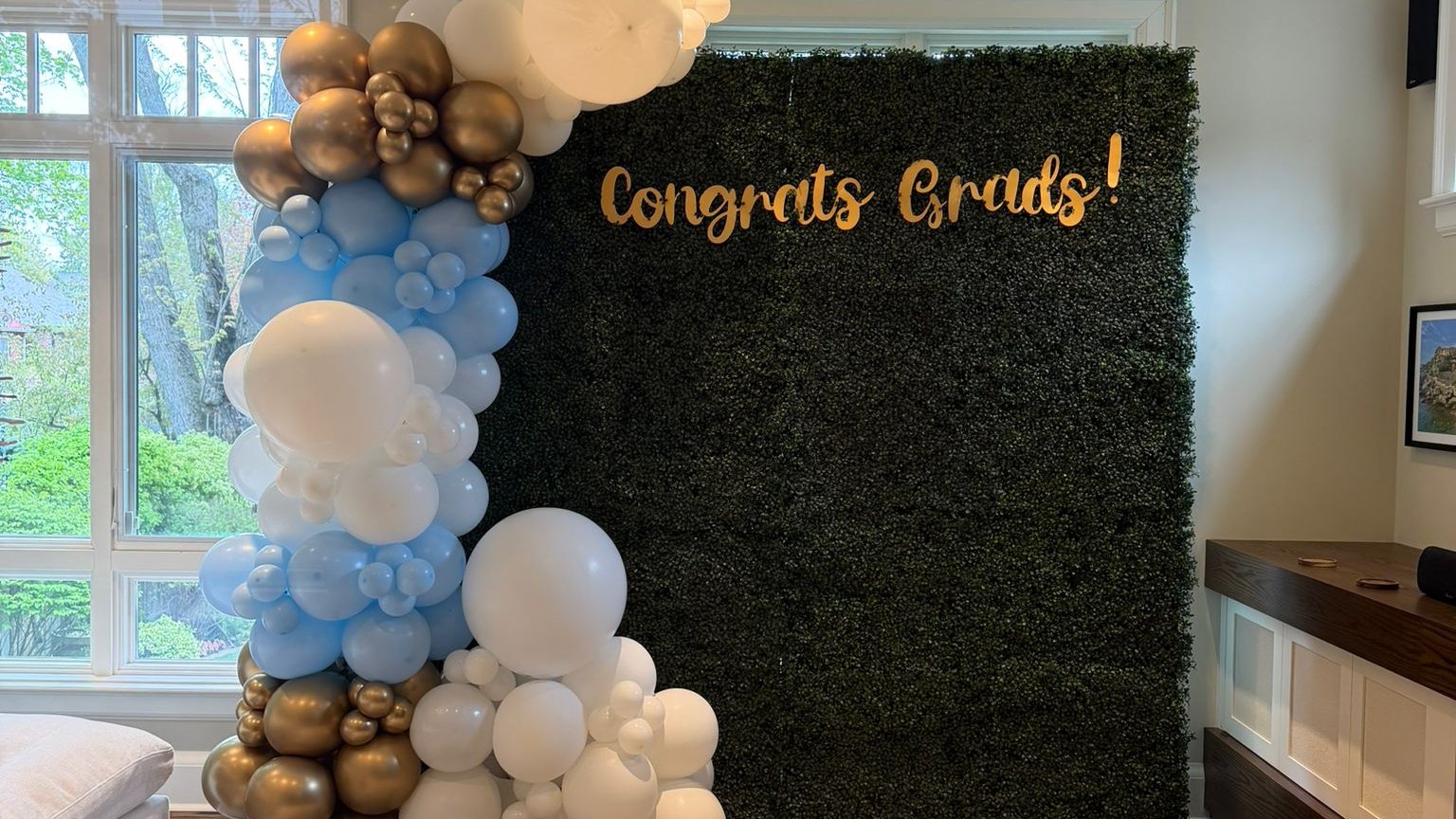 Gold script lettering mounted on a green hedge backdrop beside a blue, white and gold balloon garland