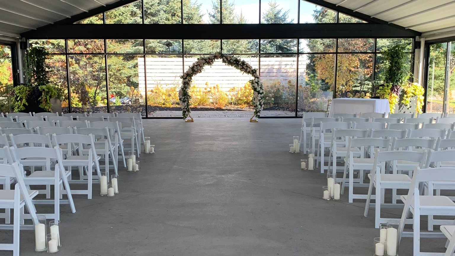 A floral arch seen the length of a glass pavilion set with rows of chairs and aisle candles