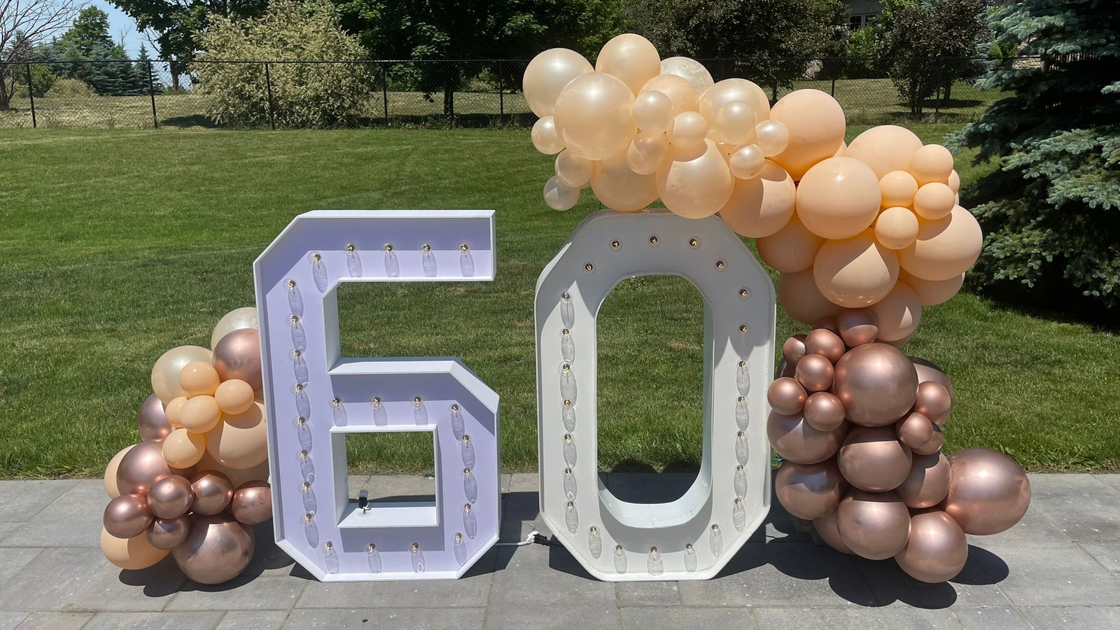 A peach, cream and rose gold balloon garland beside lit marquee numerals on a driveway