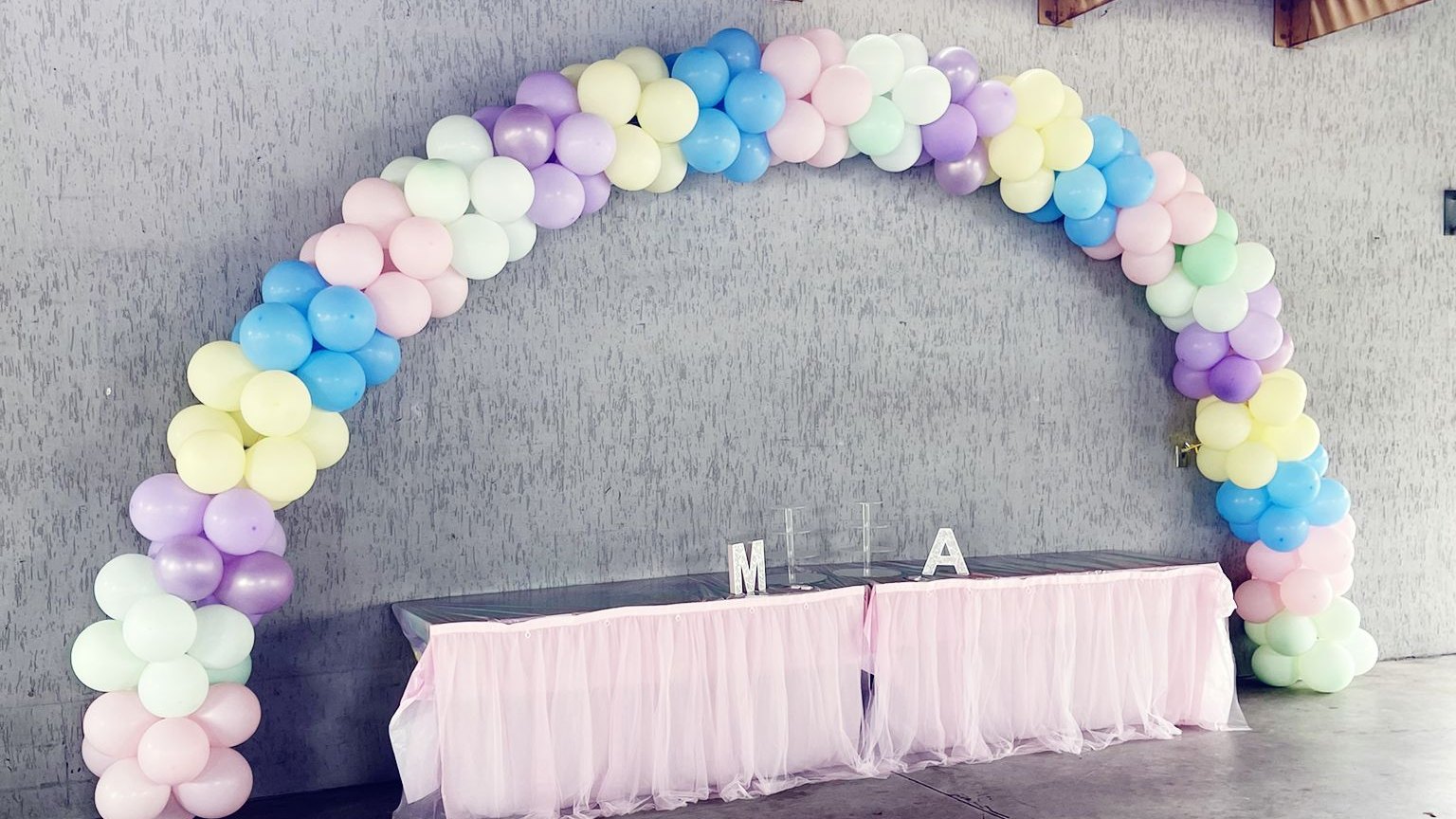 A pastel balloon arch curving over a tulle-skirted head table at a covered pavilion reception