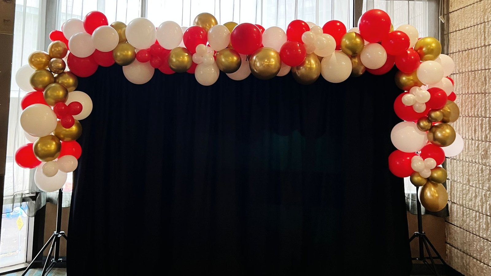 A red, gold and ivory balloon arch curving over a black fabric backdrop in a function room
